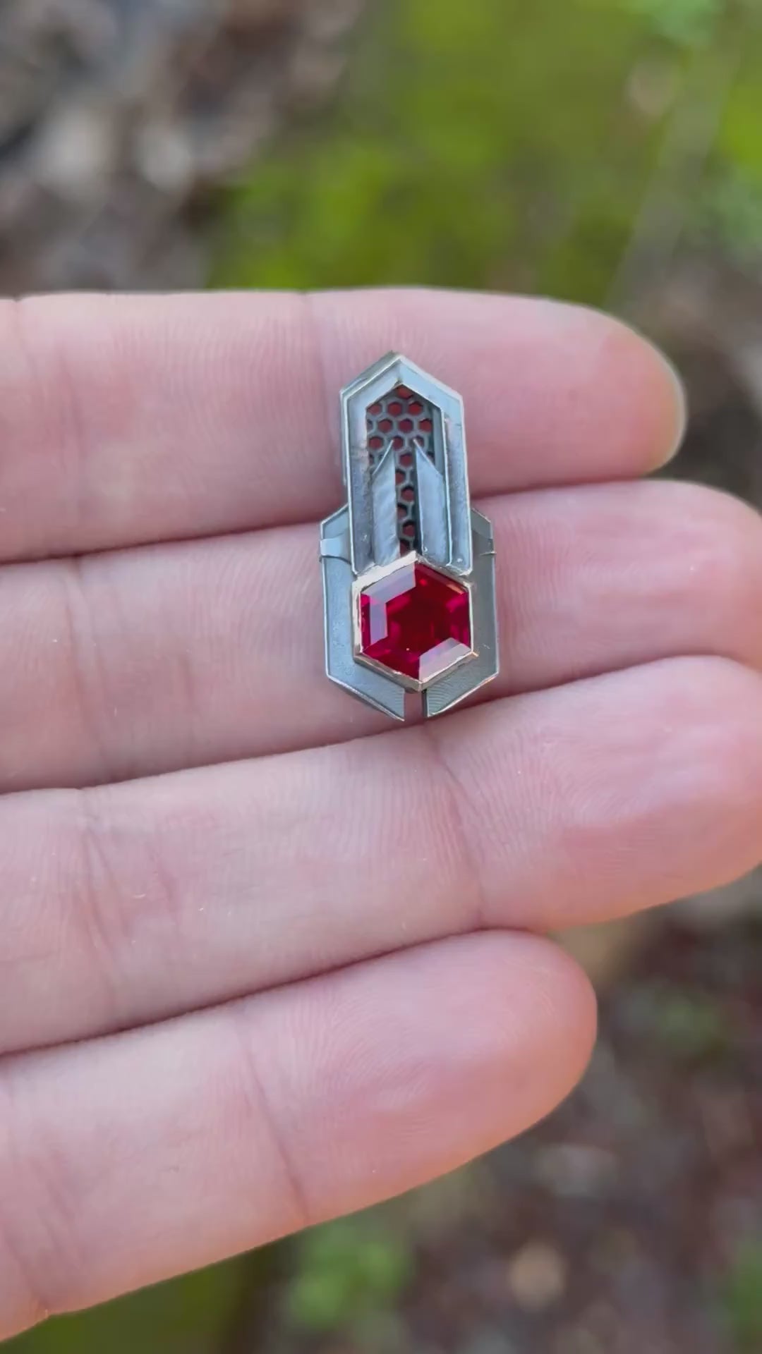 Geometric silver pendant with honeycomb details and a red gemstone, held by a hand against a grassy background.