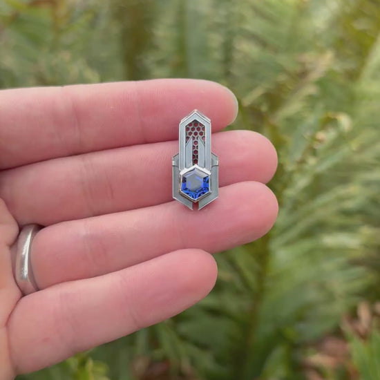 Geometric silver pendant with honeycomb details and a blue gemstone, held by a hand against a grassy background.