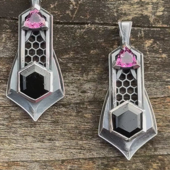 Pair of silver pendants with honeycomb details, featuring black gemstones and pink tourmalines, against a wooden background