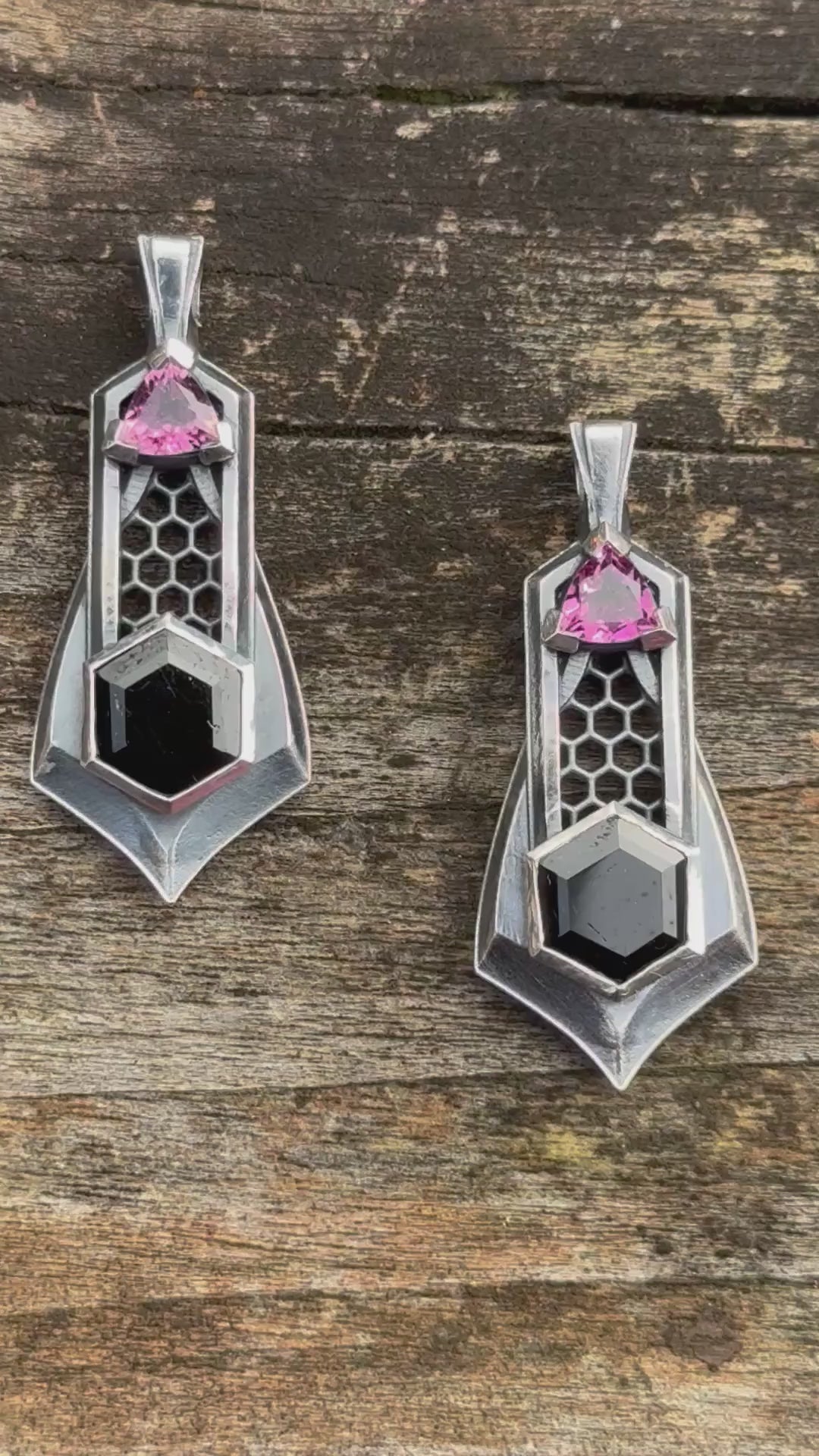 Pair of silver pendants with honeycomb details, featuring black gemstones and pink tourmalines, against a wooden background