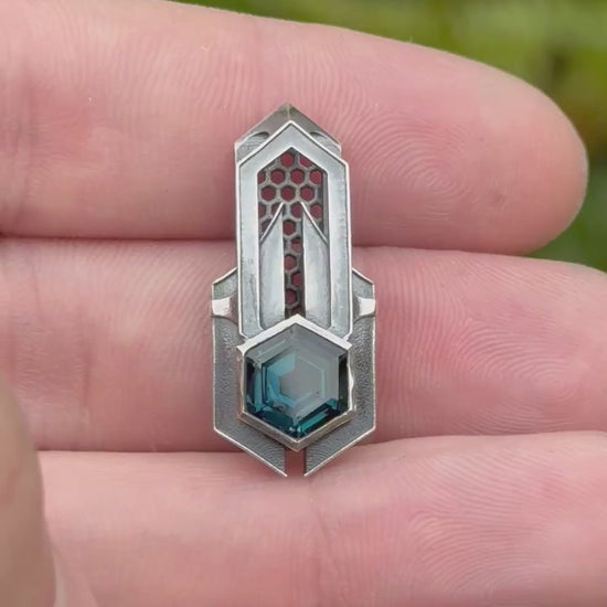 Geometric silver pendant with honeycomb details and a blue gemstone, held by a hand against a grassy background.