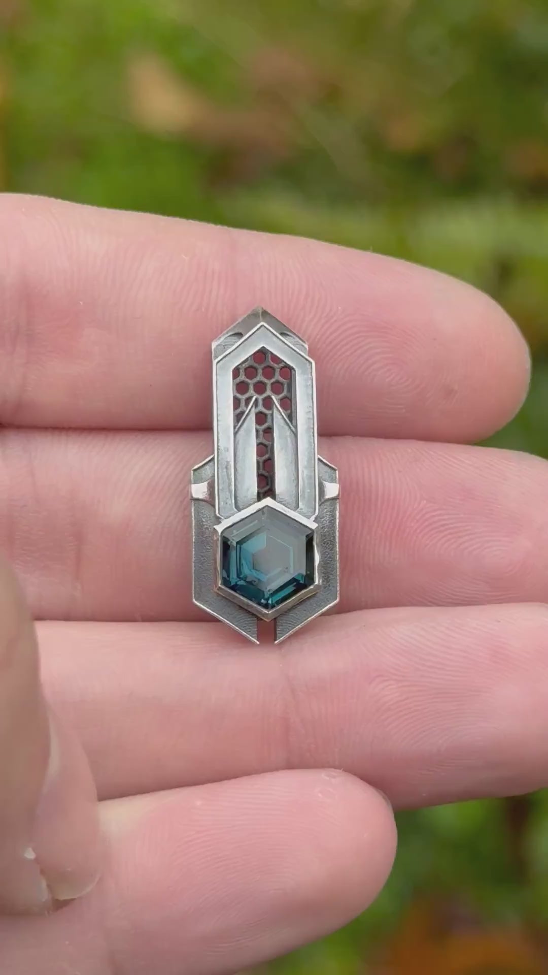 Geometric silver pendant with honeycomb details and a blue gemstone, held by a hand against a grassy background.