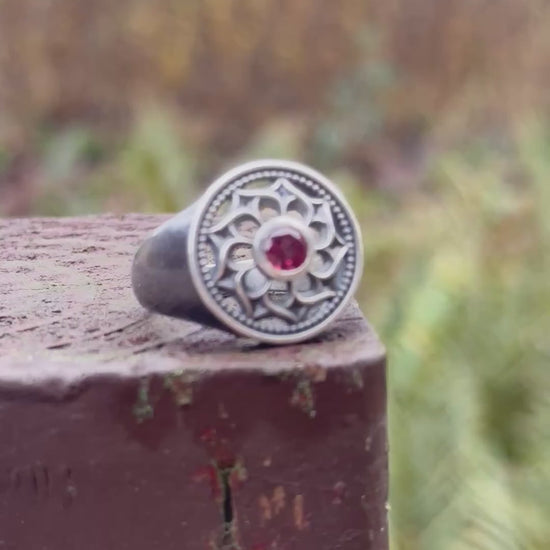 Silver ring displayed with a floral design and a red gemstone, set against a mossy outdoor scene with rocks and plants