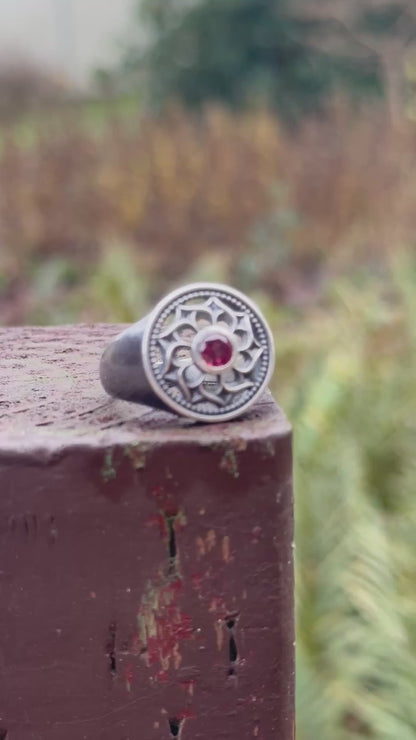 Silver ring displayed with a floral design and a red gemstone, set against a mossy outdoor scene with rocks and plants