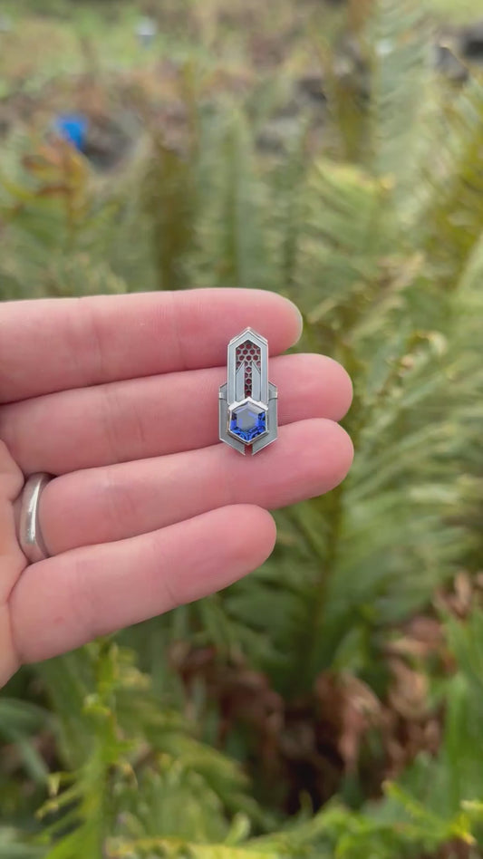 Geometric silver pendant with honeycomb details and a blue gemstone, held by a hand against a grassy background.