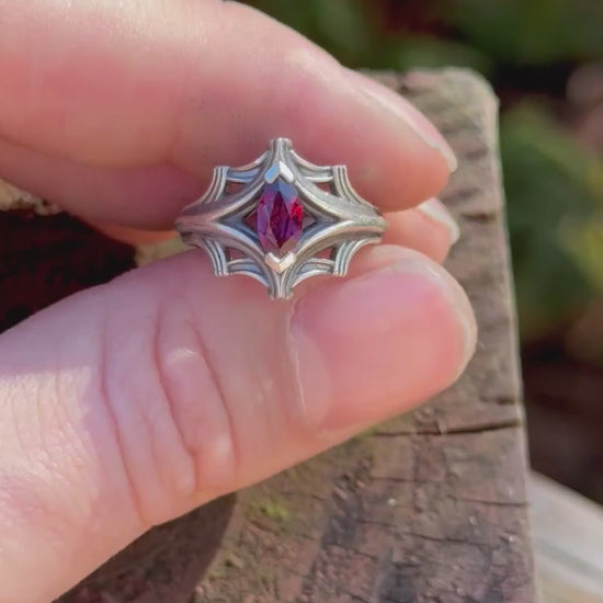 Silver ring with an ornate design featuring a large marquise-cut red gemstone, held in a hand with a fern background