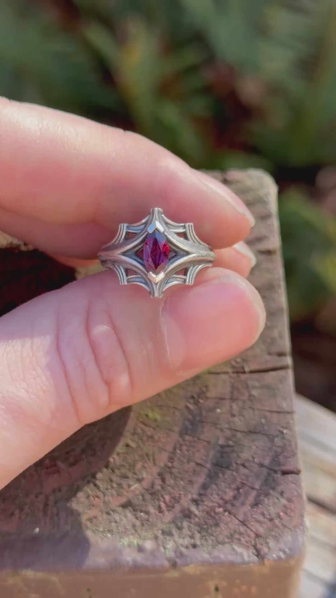 Silver ring with an ornate design featuring a large marquise-cut red gemstone, held in a hand with a fern background