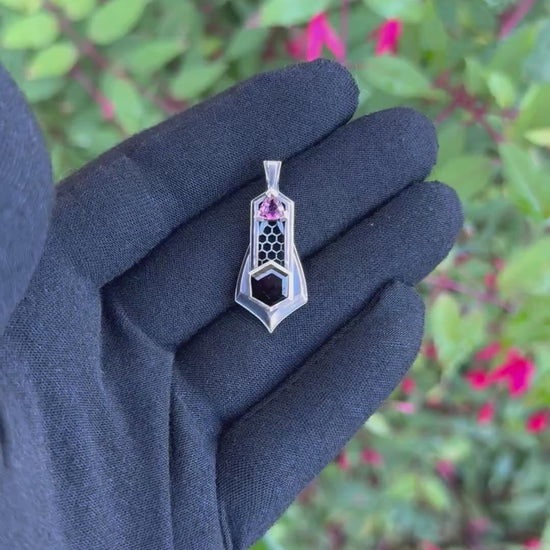 Geometric silver pendant with honeycomb details, featuring black and pink tourmaline gemstones, held by a gloved hand against a leafy background.