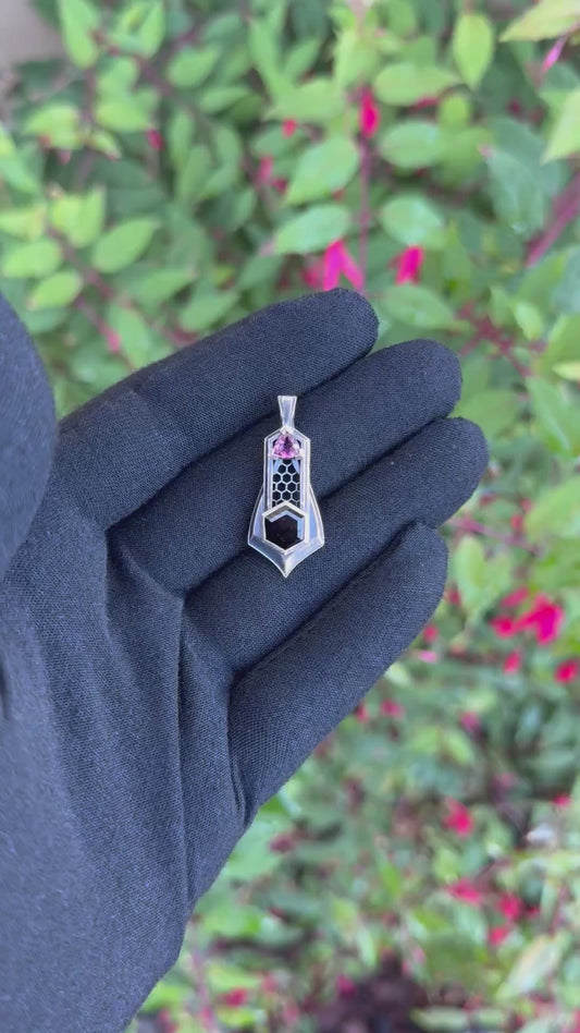 Geometric silver pendant with honeycomb details, featuring black and pink tourmaline gemstones, held by a gloved hand against a leafy background.