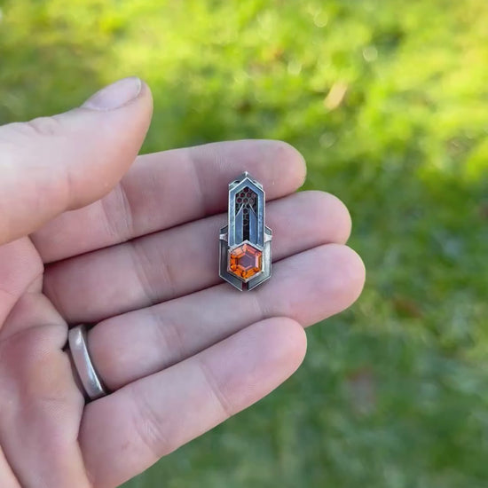 Geometric silver pendant with honeycomb details and a orange gemstone, held by a hand against a grassy background.