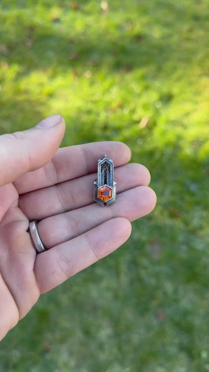 Geometric silver pendant with honeycomb details and a orange gemstone, held by a hand against a grassy background.