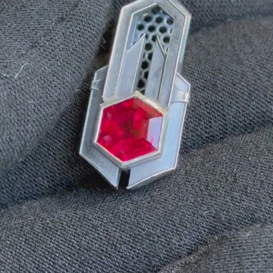 Geometric silver pendant with honeycomb details and a red gemstone, held by a gloved hand against a grassy background.