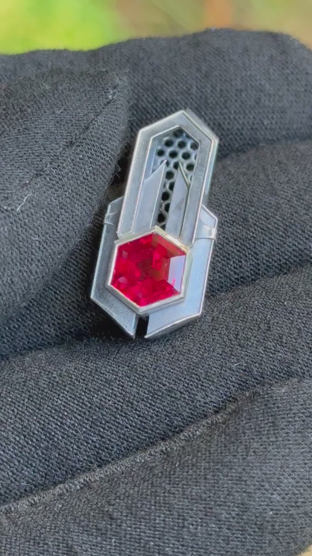 Geometric silver pendant with honeycomb details and a red gemstone, held by a gloved hand against a grassy background.