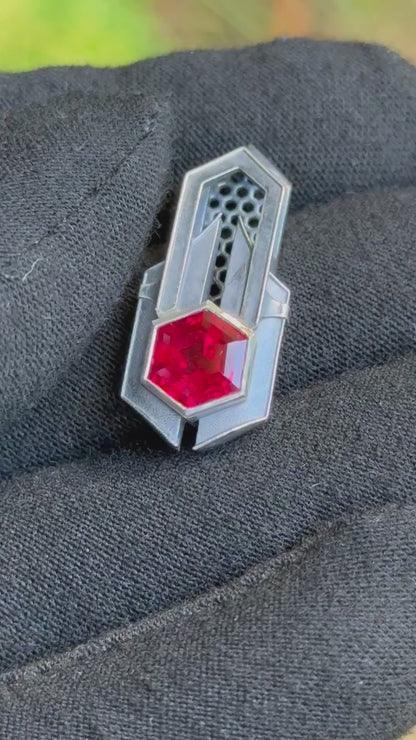 Geometric silver pendant with honeycomb details and a red gemstone, held by a gloved hand against a grassy background.