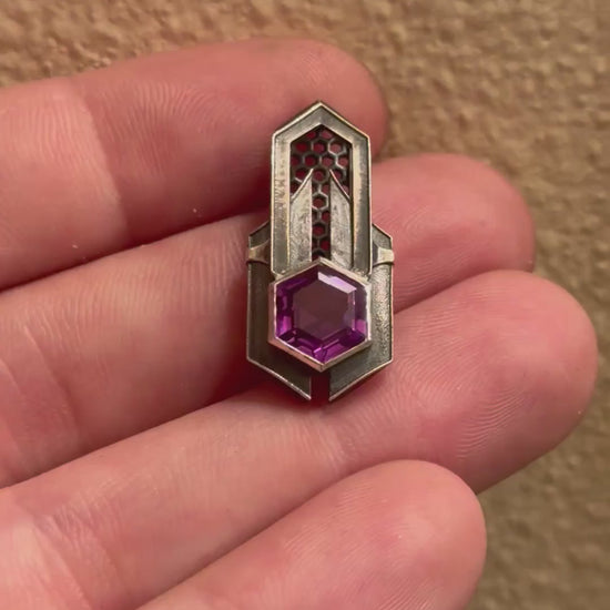 Geometric silver pendant with honeycomb details and a purple gemstone, held by a hand indoors.