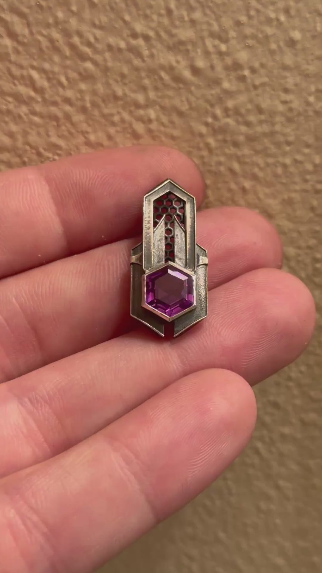 Geometric silver pendant with honeycomb details and a purple gemstone, held by a hand indoors.