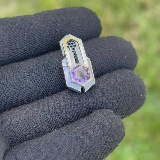 Geometric silver pendant with honeycomb details and a purple gemstone, held by a gloved hand against a grassy background.