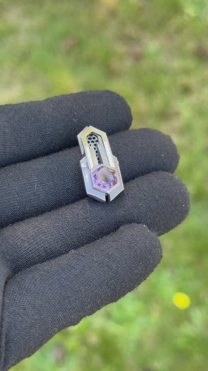 Geometric silver pendant with honeycomb details and a purple gemstone, held by a gloved hand against a grassy background.